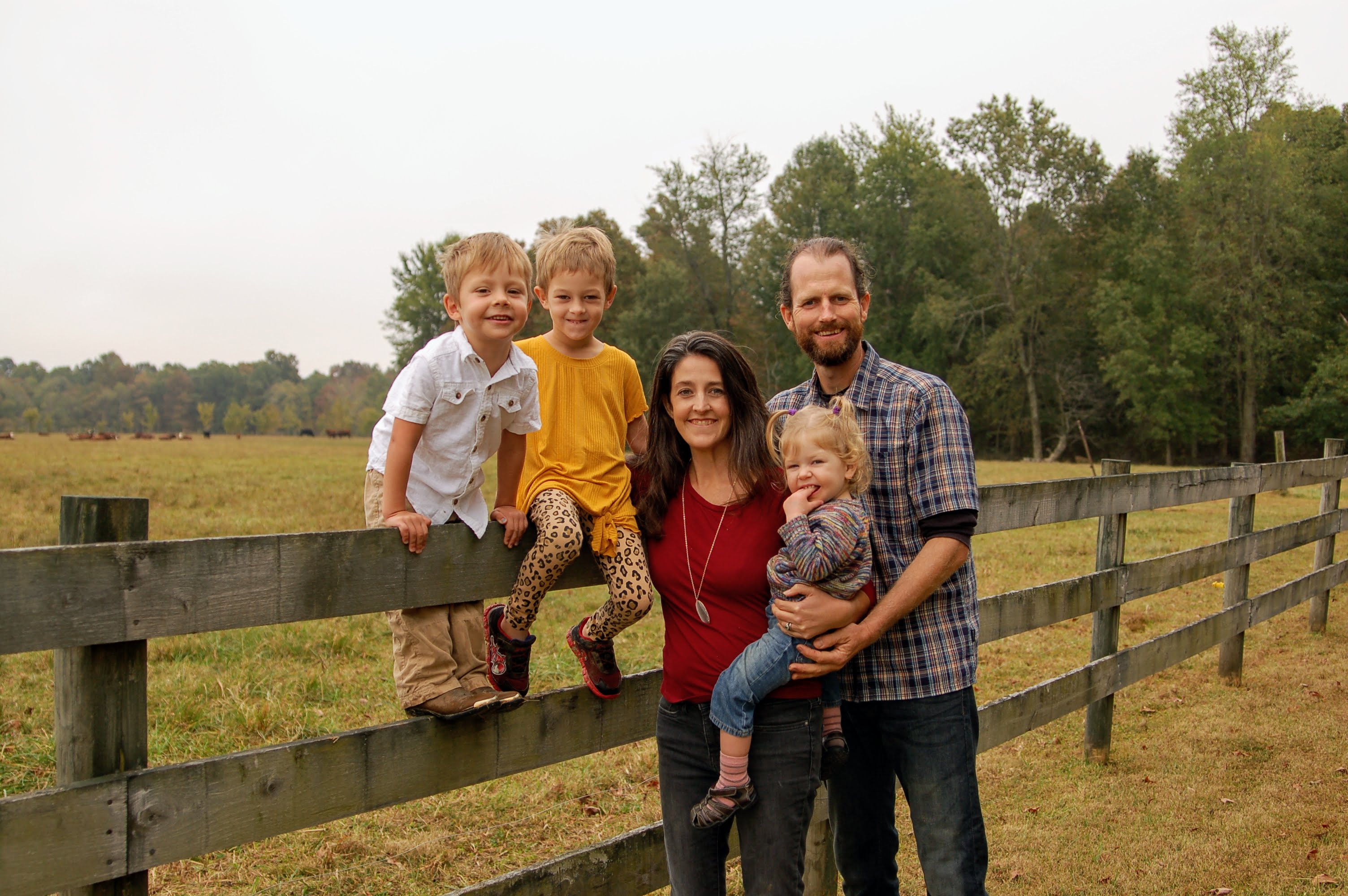 Photo of family of 5, on a fence with a field and trees in the background. Man and woman are standing, two kids are sitting on the fence, and one child is in the mother's arms.